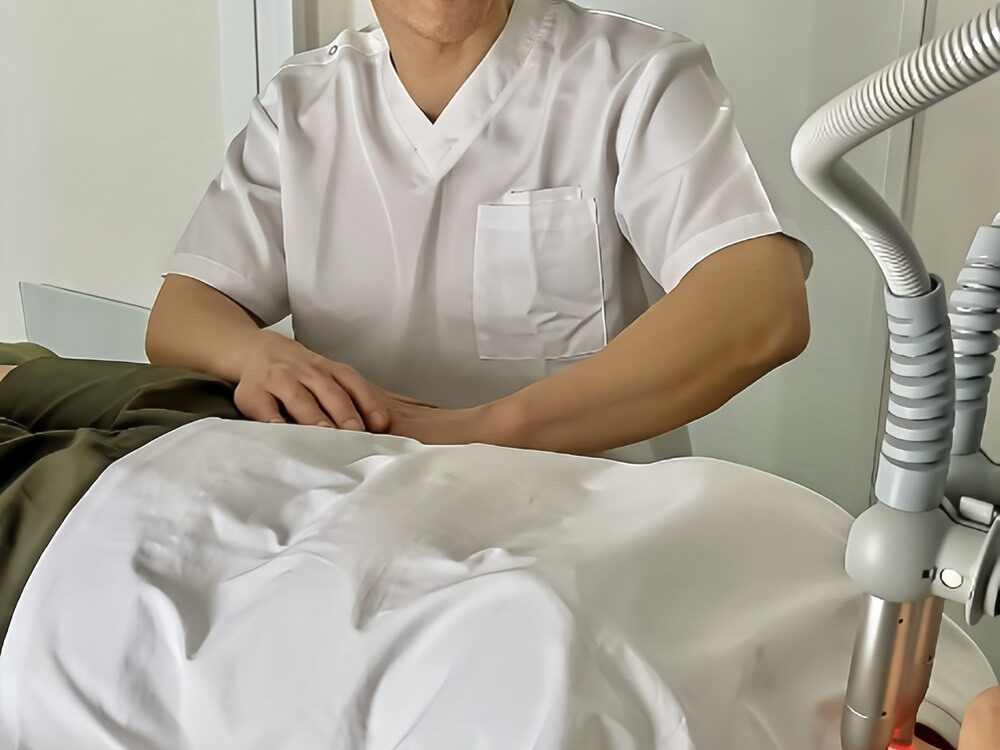 白いユニフォームを着た施術者が、横たわる患者（利用者）の腰付近に手を添えて施術を行っている様子。